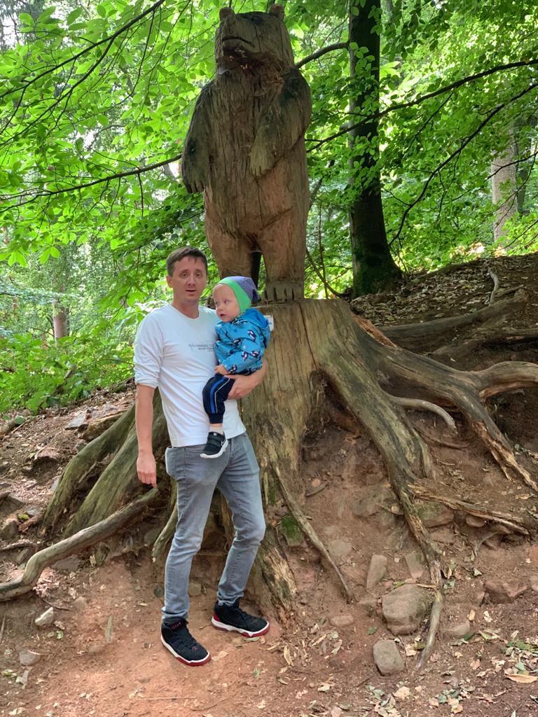 Ein Mann mit einem Kind auf dem Arm vor einer geschnitzten Bärenfigur im grünen Wald.
